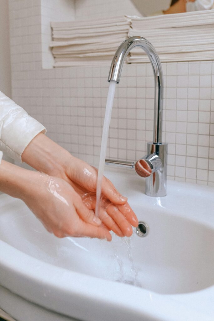 pexels-photo-3943905-3943905 Close-up of hands being washed under running water in a bathroom sink for hygiene and virus prevention.